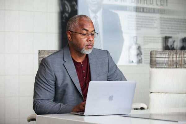 A man in a gray suit jacket works on a laptop at a white desk in a modern office space.
