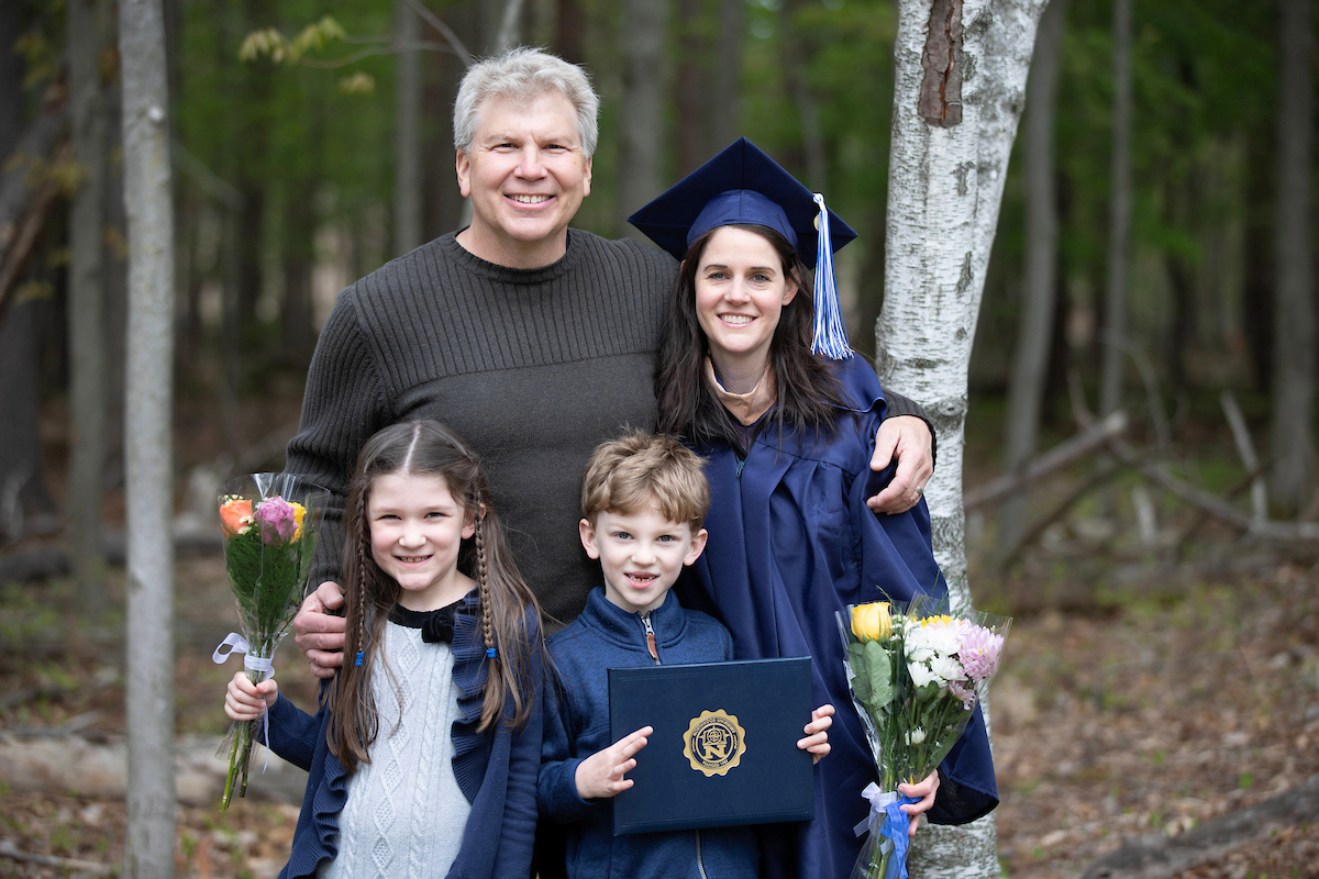 A woman in a graduation cap and gown stands with a man and two children holding flowers and a diploma in a wooded outdoor setting.