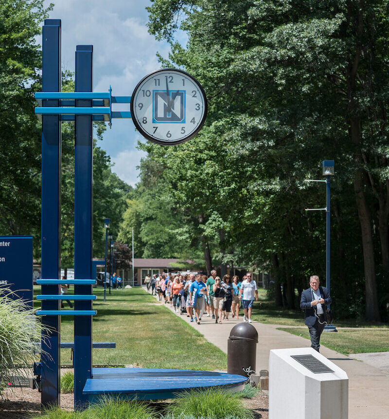 A group of people walk along a tree-lined campus path beneath a large outdoor clock displaying the time as 1:30.
