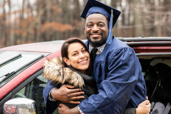 A graduate in a blue cap and gown hugs a smiling woman in a coat next to a red car on a rainy day.