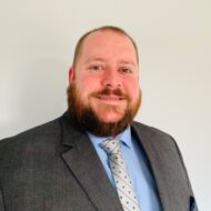 A man with a beard wearing a gray suit, light blue shirt, and patterned tie stands in front of a plain light-colored background, smiling at the camera.