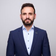A man with short brown hair and a beard, wearing a navy blue suit and light blue striped shirt, stands against a plain light gray background.