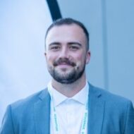 A man with short hair and a beard, wearing a blue suit jacket, white shirt, and conference lanyard, stands in front of a light blue background, smiling at the camera.