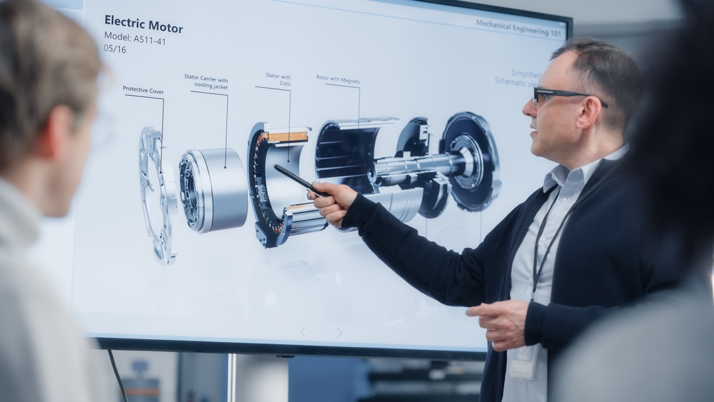 Man in glasses points to a diagram of an electric motor on a large screen during a technical presentation to a small group.