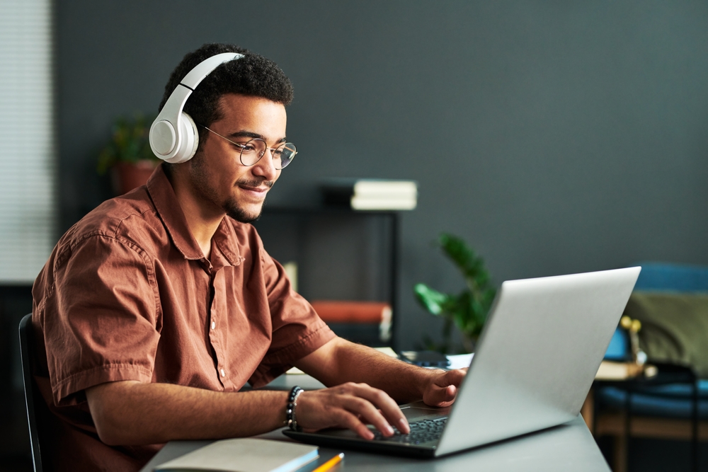 A man wearing headphones and glasses sits at a desk using a laptop in a modern office setting.