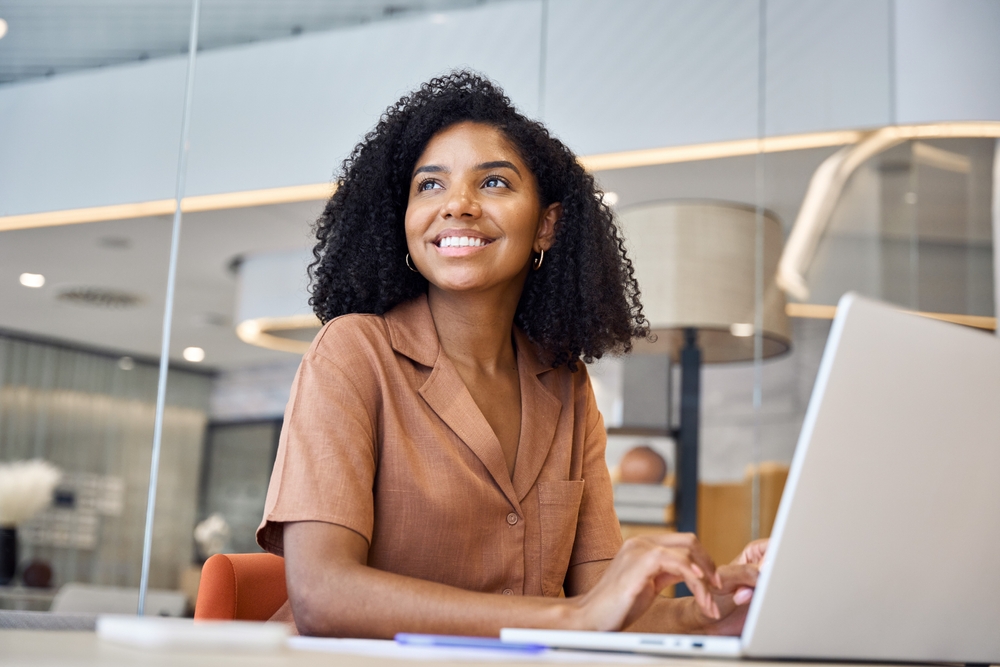 A woman with curly hair and a brown shirt sits at a desk, smiling while typing on a laptop in a modern office setting.