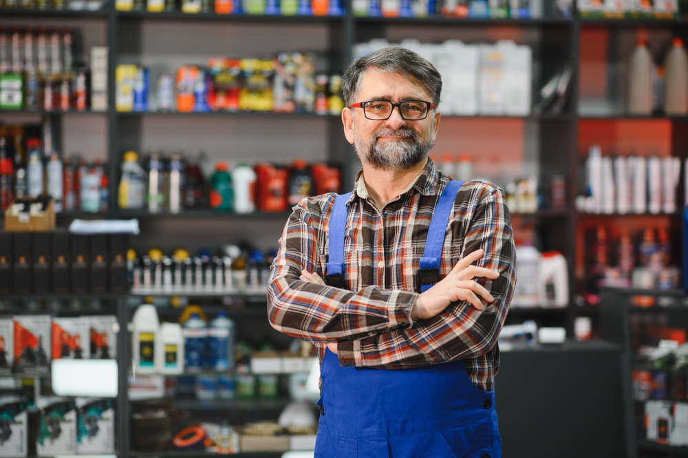 A man in a plaid shirt and blue overalls stands with folded arms in front of shelves filled with automotive products in a store.