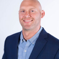 A bald man wearing a dark blue blazer and light blue shirt smiles at the camera against a plain white background.