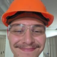 A person wearing an orange hard hat, safety glasses, and a gray shirt smiles in a hallway with lockers in the background.