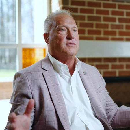 A middle-aged man with short hair in a white dress shirt and plaid blazer gestures while speaking in a customer testimonial carousel.