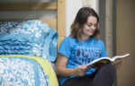 A young woman in a blue "Timberwolves" t-shirt sits on the floor by a bed with blue floral bedding, reading a book and smiling.