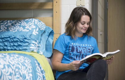 A young woman in a blue "Timberwolves" t-shirt sits on the floor by a bed with blue floral bedding, reading a book and smiling.