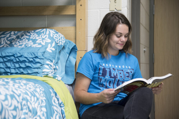 A young woman in a blue "Timberwolves" t-shirt sits on the floor by a bed with blue floral bedding, reading a book and smiling.