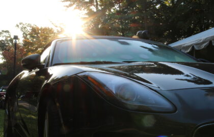 A close-up view of a black sports car parked outdoors with sunlight shining through trees in the background.