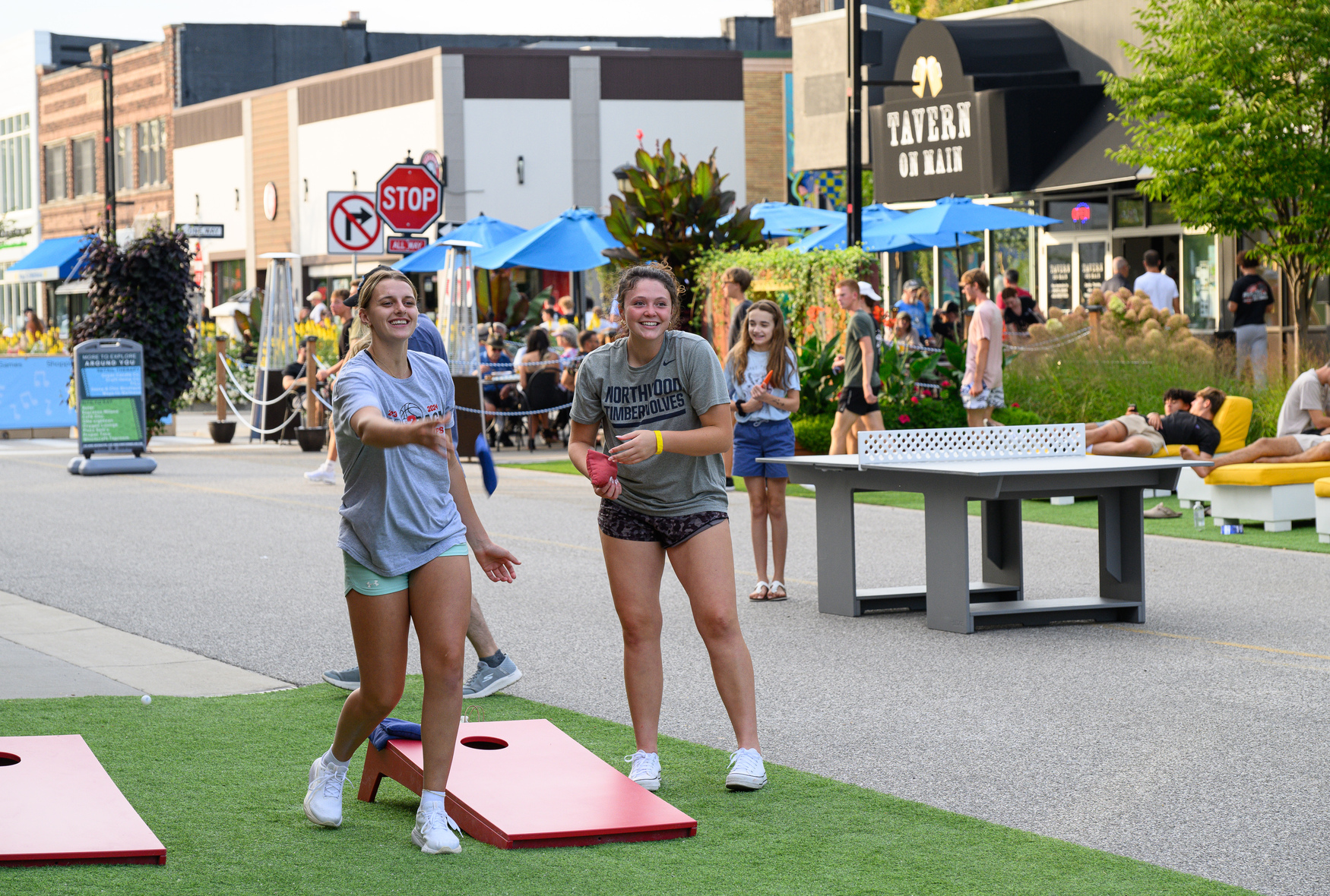 Two young women play cornhole on artificial grass in a lively shopping and dining area near Midland College, with people and tables bustling in the background.