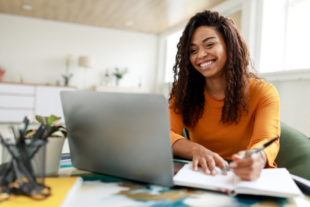 Portrait of smiling woman sitting at desk, using laptop and writing in notebook.