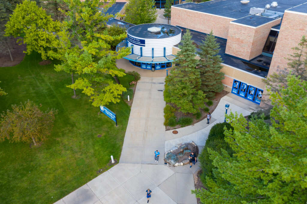 Aerial view of one of the best college campuses, showing a building entrance surrounded by trees, with several people walking on paved paths and a blue welcome banner near the doors.