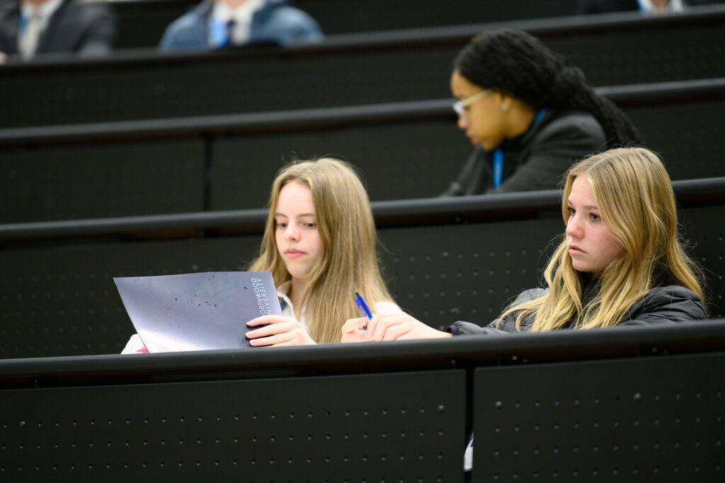 Three students sit in a lecture hall on one of the best college campuses; two in the foreground focus on notes and a booklet, while one in the background looks ahead.