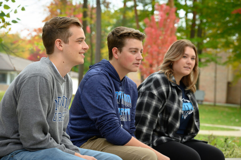 Three young adults sit outdoors on a bench, wearing casual clothing and sweatshirts, enjoying one of the best college campuses with vibrant autumn trees and a building in the background.