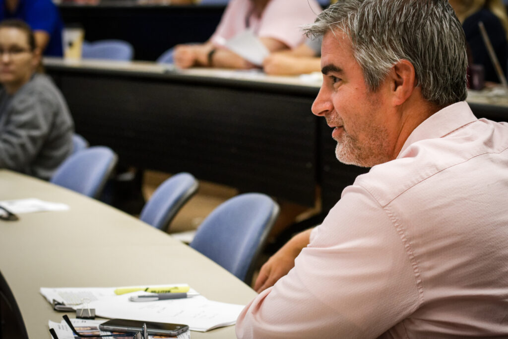 A man in a pink shirt, possibly a Doctor of Business Administration student, sits at a desk in a lecture hall, looking to the side. Papers, highlighters, and pens are on the desk, with others seated in the background.
