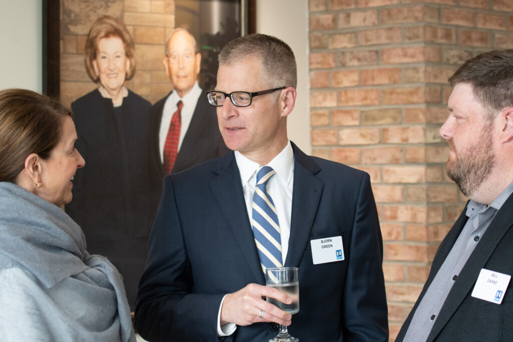 Three people in business attire, including a Doctor of Business Administration, converse indoors. Two wear name tags and one man holds a glass of water. A framed photo of an elderly couple hangs on the wall behind them.