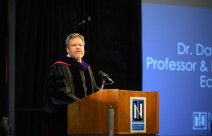 A man in academic regalia stands at a podium, speaking at an event. A presentation slide behind him displays "Dr. D..." and "Doctor of Business Administration, Professor &..." at Northwood University.