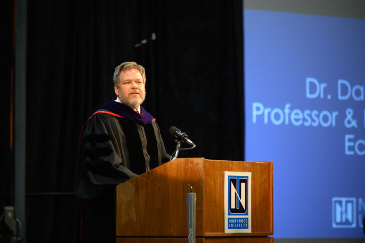 A man in academic regalia stands at a podium, speaking at an event. A presentation slide behind him displays "Dr. D..." and "Doctor of Business Administration, Professor &..." at Northwood University.