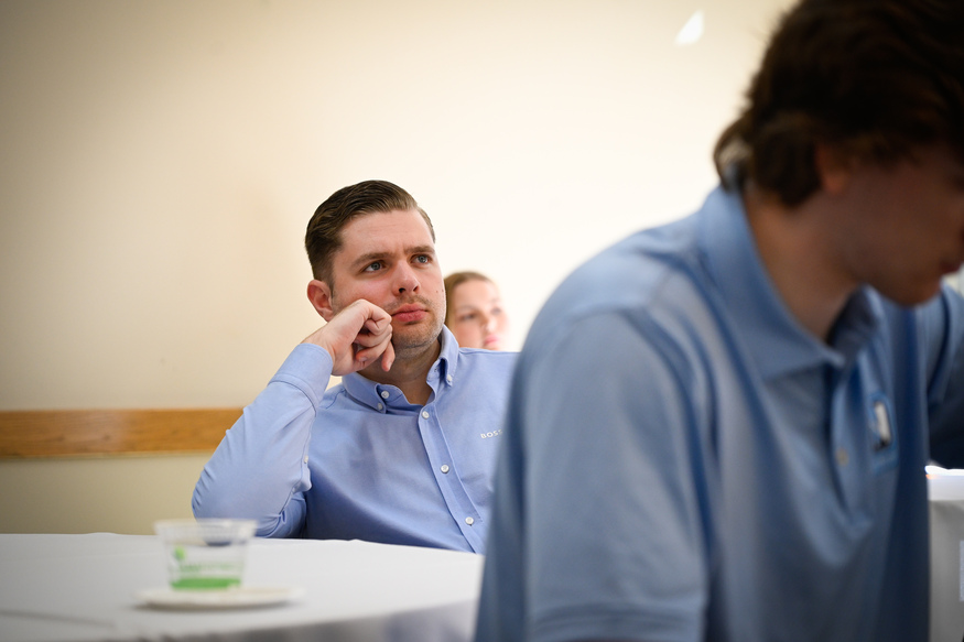 A man in a blue button-up shirt sits at a table, resting his chin on his hand and looking ahead, perhaps contemplating business schools, while another person in the foreground is turned away.