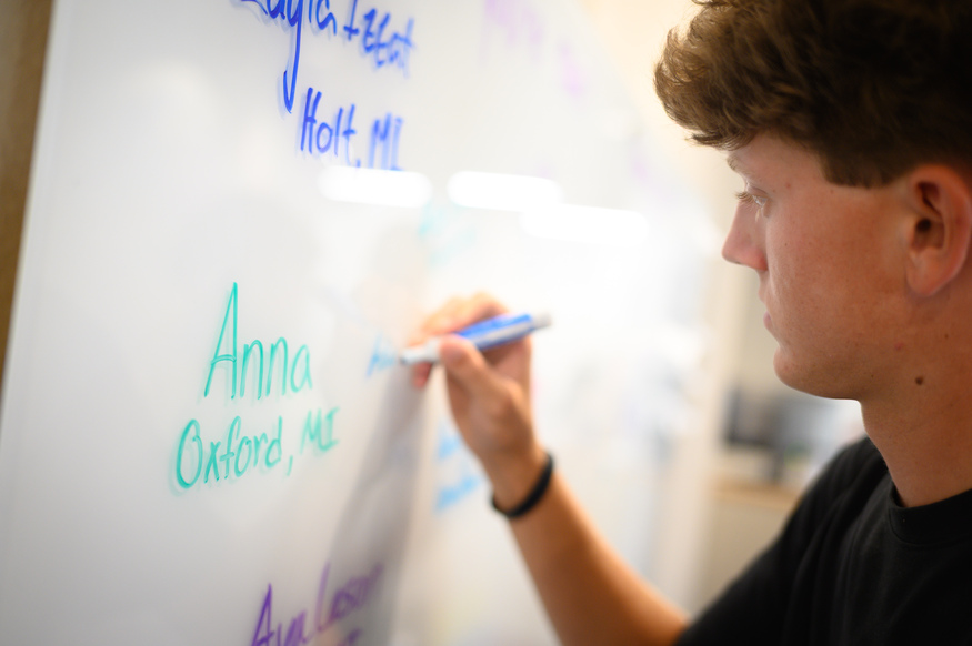 A person writes names and locations, including "Anna, Oxford, MI," and notable business schools on a whiteboard with a blue marker.