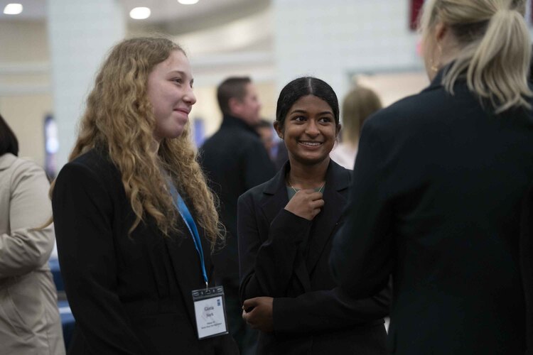 Two young women in business attire, possibly networking at a business schools event, smile and chat with another person indoors; one wears a name badge.