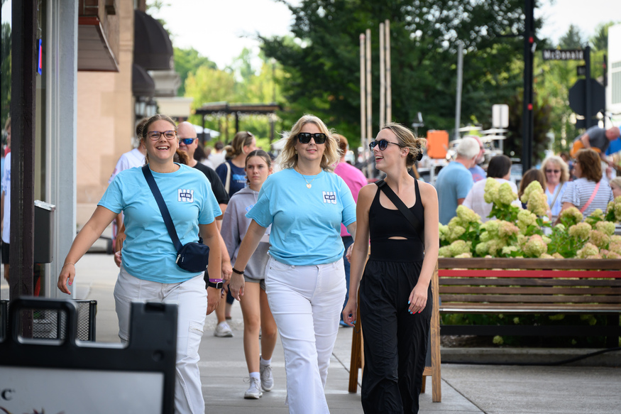 Three women walk down a busy sidewalk near what appears to be business schools; two wear matching blue t-shirts and white pants, while the third wears a black tank top and pants. Other people are visible in the background.