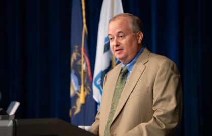 A man in a tan suit and green tie speaks at a podium, possibly addressing an audience from leading business schools, with flags and a dark blue curtain in the background.