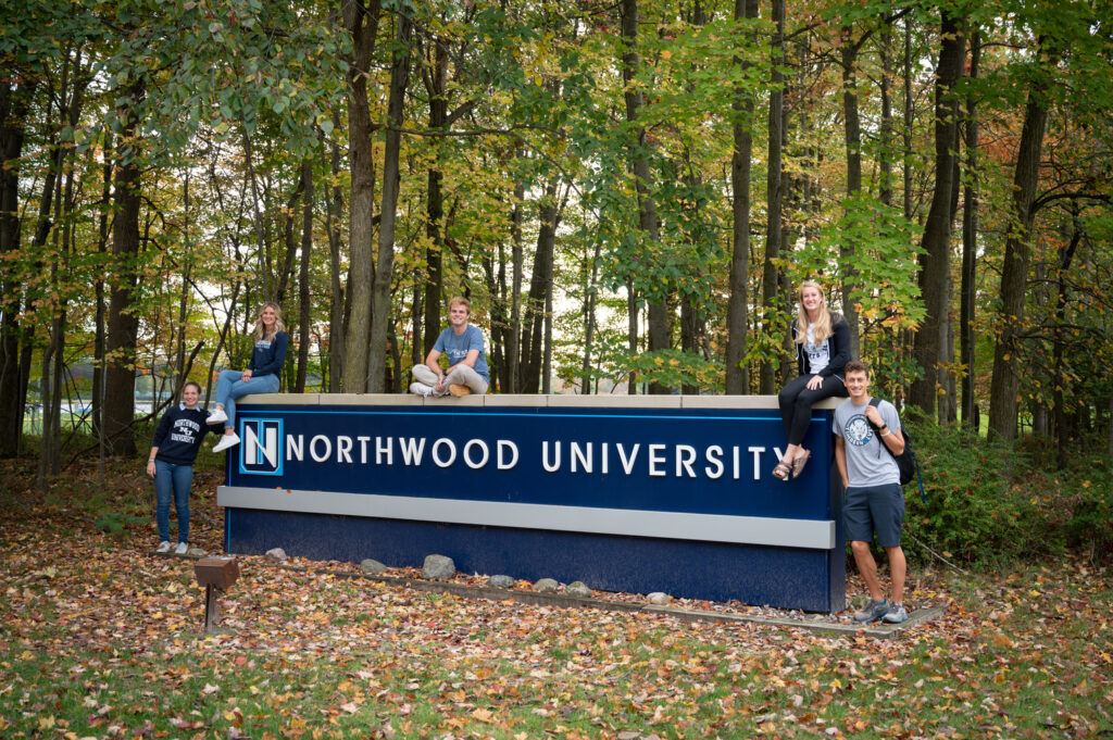 Five people pose around and on top of a large Northwood University sign set in a wooded area with autumn foliage, highlighting the vibrant community at one of the leading business schools.