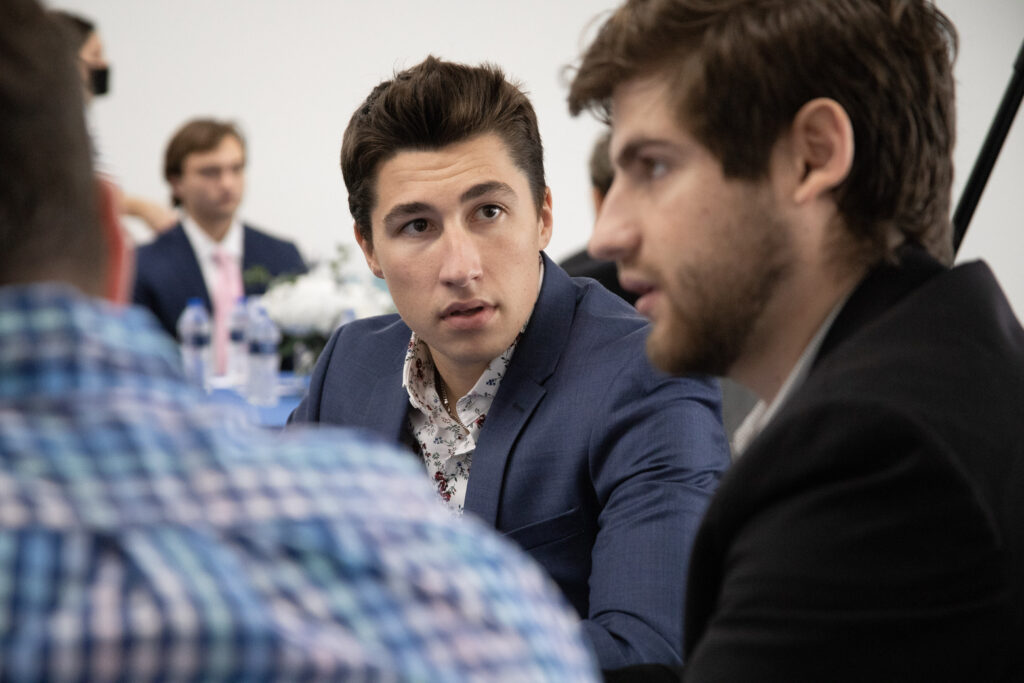 Two men in suits have a serious conversation at a table, possibly discussing strategies relevant to business schools, while other people are blurred in the background.