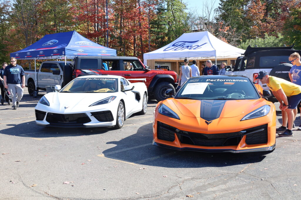 Two Chevrolet Corvette sports cars, one white and one orange, parked at an outdoor car show with people and Ford-branded tents in the background—an exciting scene for motorsports jobs enthusiasts.