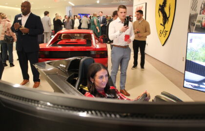 A woman uses a racing simulator in the foreground, while people gather and talk near a red Ferrari in a showroom setting, highlighting the excitement of motorsports jobs.
