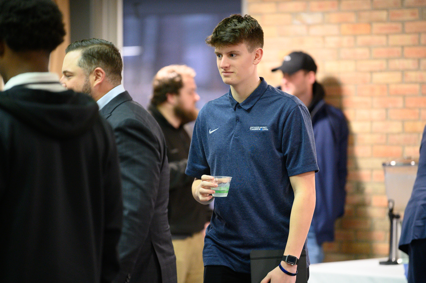 A young man wearing a blue polo shirt holds a drink and a folder while standing among peers at an indoor event for one of the Best Undergraduate Business Schools.