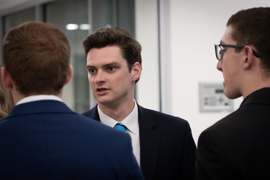 Three men in suits have a conversation indoors, perhaps discussing opportunities at the Best Undergraduate Business Schools, with one man in the center facing the camera and speaking.