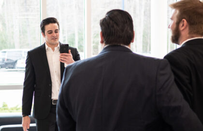 In a bright office lobby, a man in a black suit, possibly discussing the Best Undergraduate Business Schools, holds a smartphone and speaks to two other suited men with their backs to the camera.