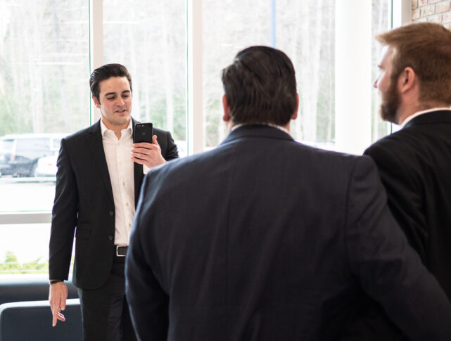 In a bright office lobby, a man in a black suit, possibly discussing the Best Undergraduate Business Schools, holds a smartphone and speaks to two other suited men with their backs to the camera.