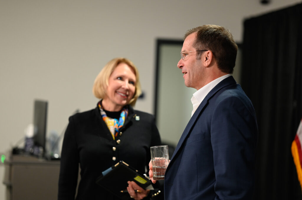 A woman holding a notebook and a man holding a glass of water are having a conversation in an indoor setting, possibly discussing opportunities at universities in Michigan.