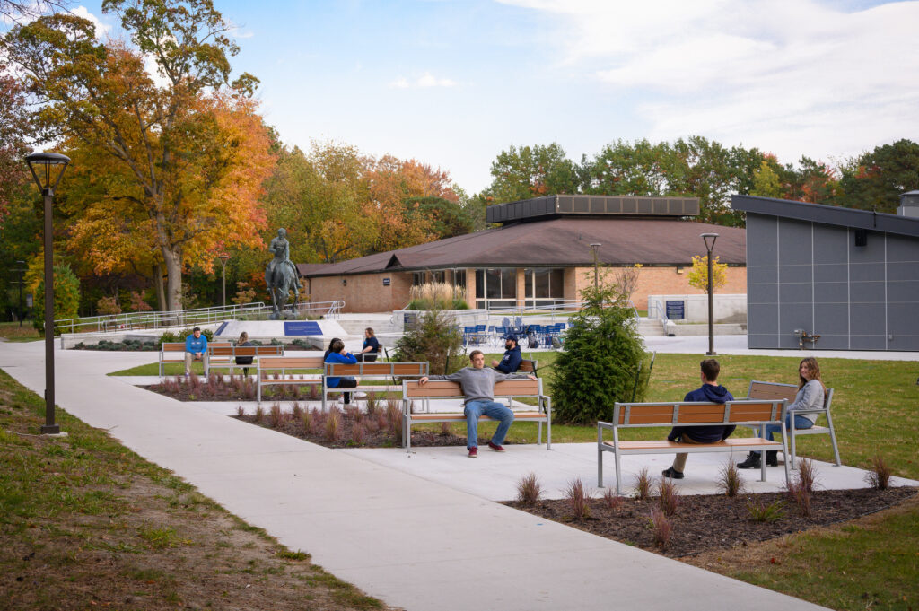 People sit on benches along a paved path in an outdoor area near a modern building, with trees showing autumn colors—a tranquil scene often found at universities in Michigan.