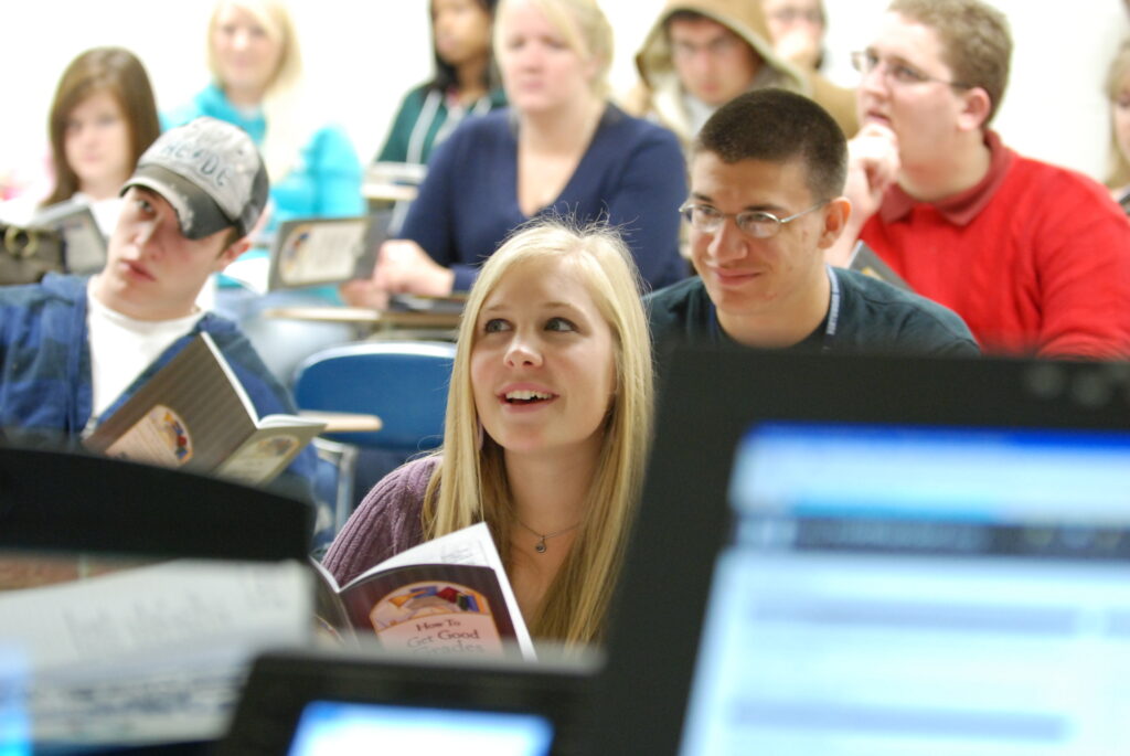 Students sit in a classroom at one of the universities in Michigan, some holding books and notebooks, attentively listening and looking toward the front of the room.