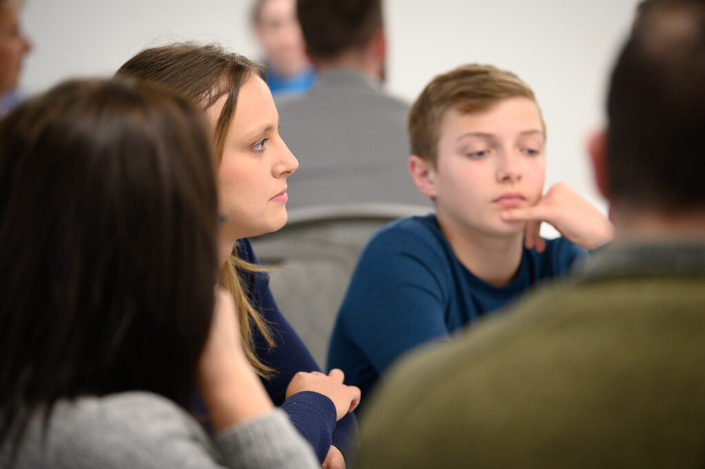 A group of people sit together at a table, focusing on a discussion about universities in Michigan. A woman and a boy are clearly visible, both looking attentive.