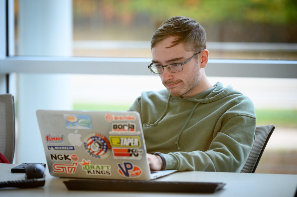 A person wearing glasses and a green hoodie types on a sticker-covered laptop, possibly researching universities in Michigan, while seated at a desk near a large window.