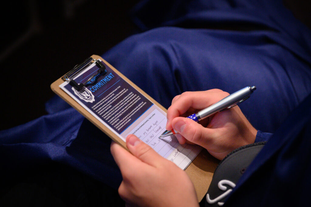 A person with a Bachelor Degree holds a clipboard and fills out a form labeled "Commitment" with a pen, wearing a dark blue jacket and cap.