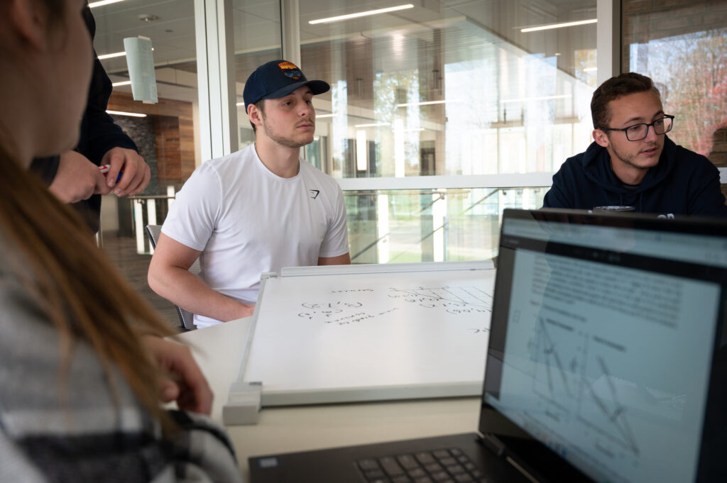 Four people with a Bachelor Degree sit around a table, collaborating in a bright indoor space with a whiteboard full of diagrams and a laptop displaying a document.