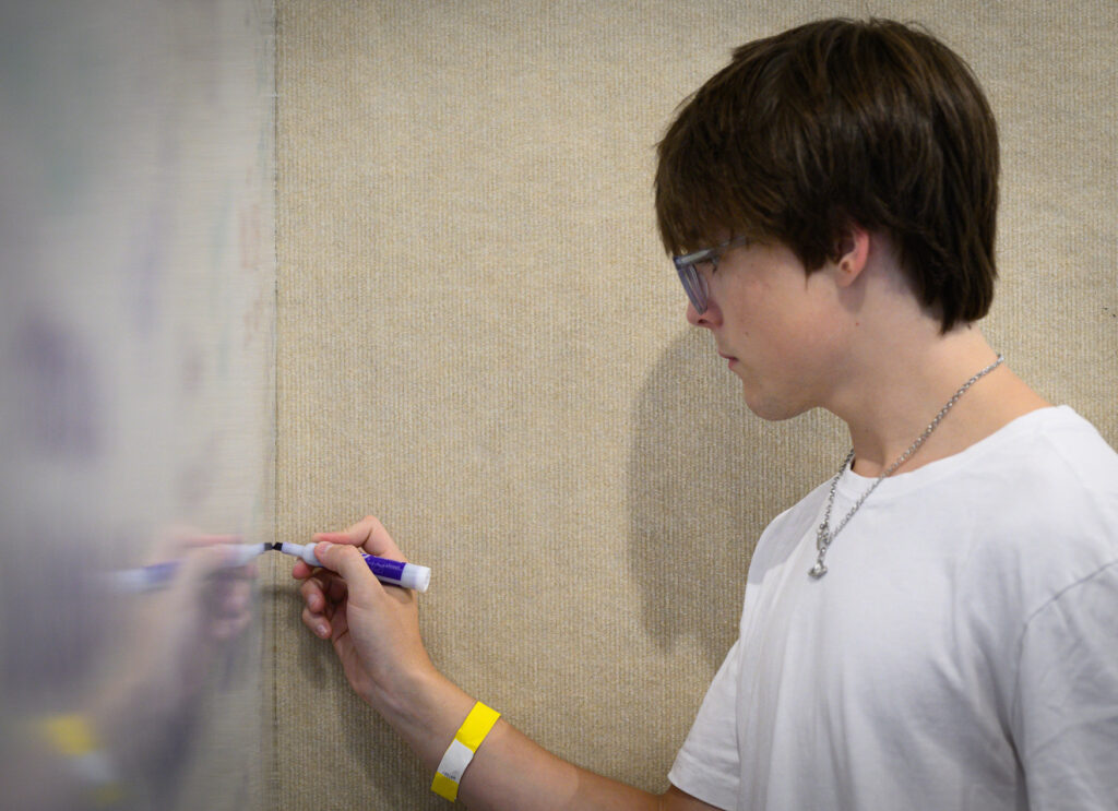 A young person with glasses and a yellow wristband, possibly pursuing a Bachelor Degree, writes on a whiteboard with a blue marker, wearing a white t-shirt and chain necklace.
