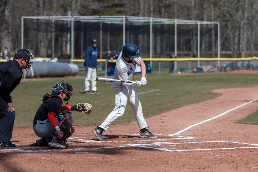 A baseball player in a white uniform swings at a pitch during a game, as the scene captures the excitement of sports marketing with a catcher, umpire, and other players visible in the background.