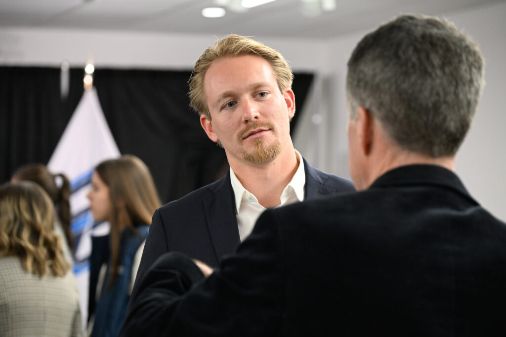 A man in a suit listens to another person speaking at an indoor sports marketing event, with several people and a flag in the background.